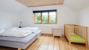 A bedroom with a wooden ceiling, a large window with a view of greenery, a bed with white bedding, a wooden crib, and a small dresser.