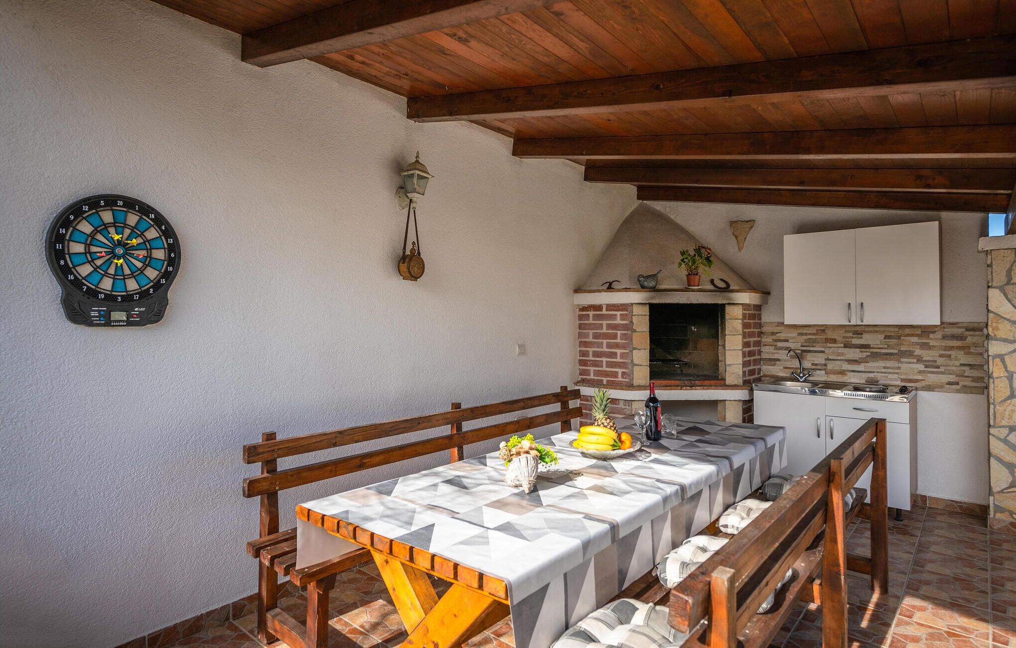 A dining area with a table set for two, a dartboard, a hanging lantern, and a kitchen with a brick oven.