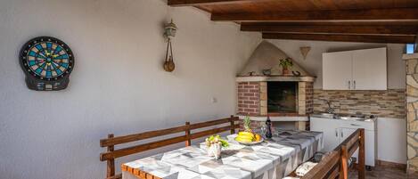 A dining area with a table set for two, a dartboard, a hanging lantern, and a kitchen with a brick oven.
