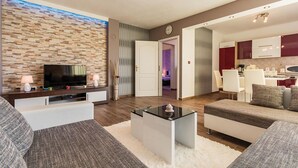 A modern living room with a flat-screen TV, a coffee table, and a sofa. There is a kitchen area with white cabinets and a dining table.