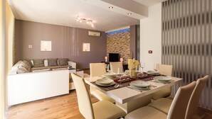 A modern dining area with a central table set for six, surrounded by beige chairs. A stone wall and a television are visible in the background.