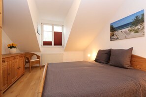 A bedroom with a large bed, a wooden dresser, a wicker chair, a radiator, a window with blinds, and a beach scene painting on the wall.