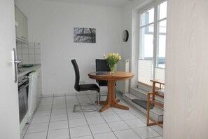 A compact kitchen with white cabinets, a black chair, a wooden table with a vase of flowers, and a view through a window.