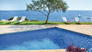 A pool with a view of the sea and boats, surrounded by lounge chairs and a tree.