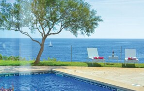 A pool area with two lounge chairs and a table, overlooking a clear blue sea and a sailboat. - Gorgeous home in Porto Colom (Porto Colom)