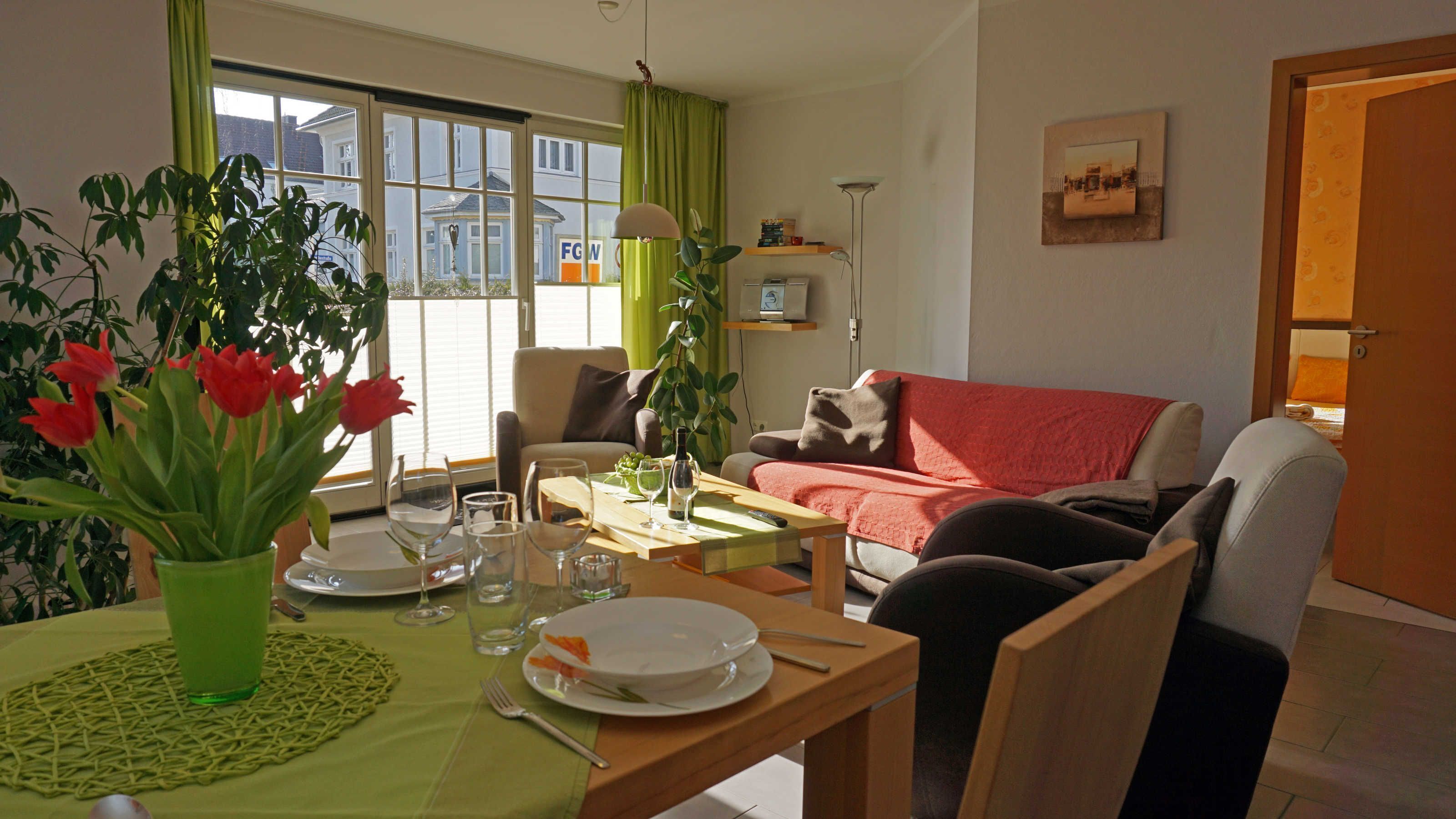 Ein Wohnzimmer mit Essbereich für zwei Personen, einer Couch, einem Sessel und einem Blumentopf mit roten Blüten.