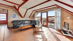Un vaste espace de vie comprenant un canapé en cuir noir, une table basse en bois et un fauteuil à bascule. Un téléviseur est posé sur un meuble, et une grande fenêtre est agrémentée de rideaux.