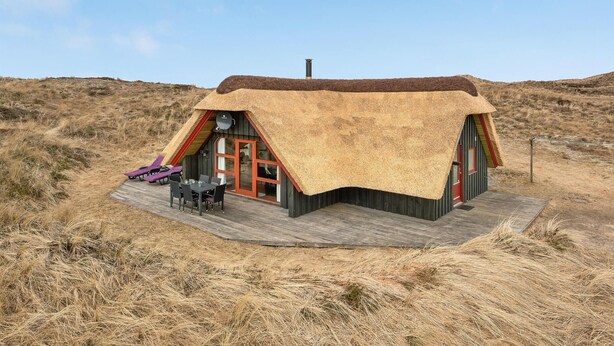 Une maison à toit de chaume avec une terrasse en bois, un coin repas avec des chaises et une antenne parabolique.