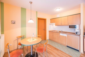 A compact kitchen with wooden cabinets, a microwave, and a dishwasher. There is a dining table with four chairs and a hanging light fixture.