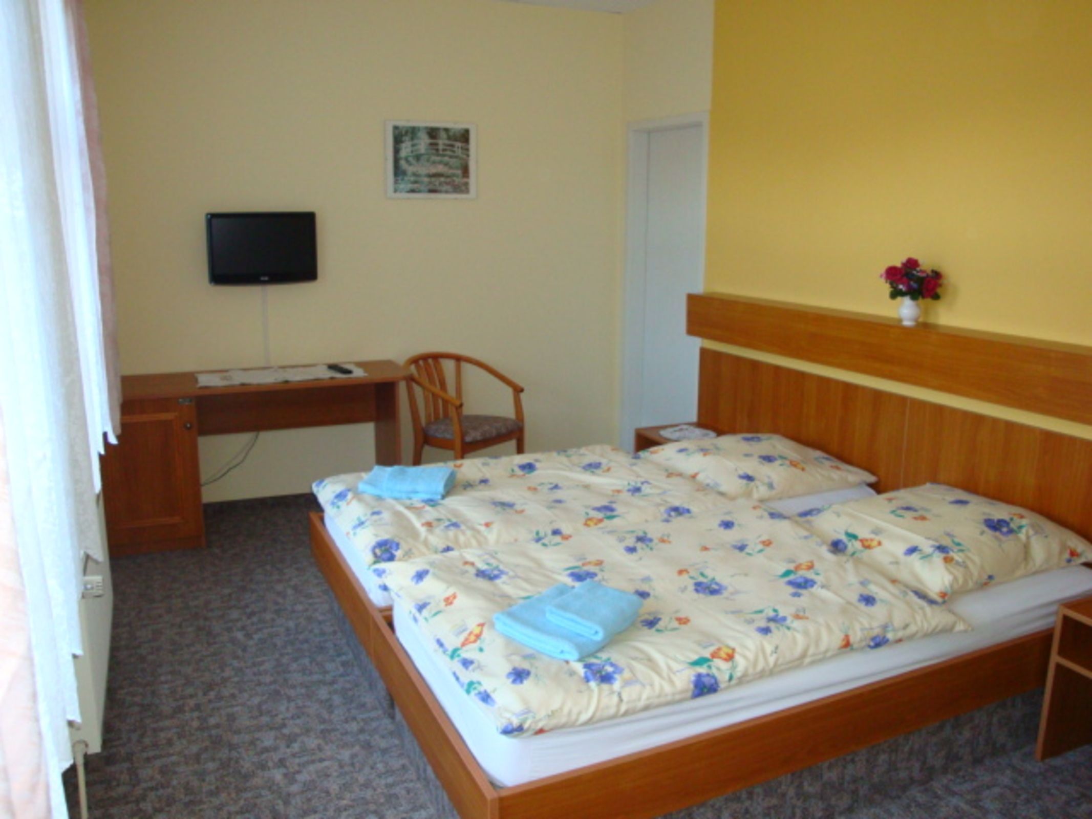 Un dormitorio con cama, escritorio, silla, televisor y un pequeño jarrón con flores.