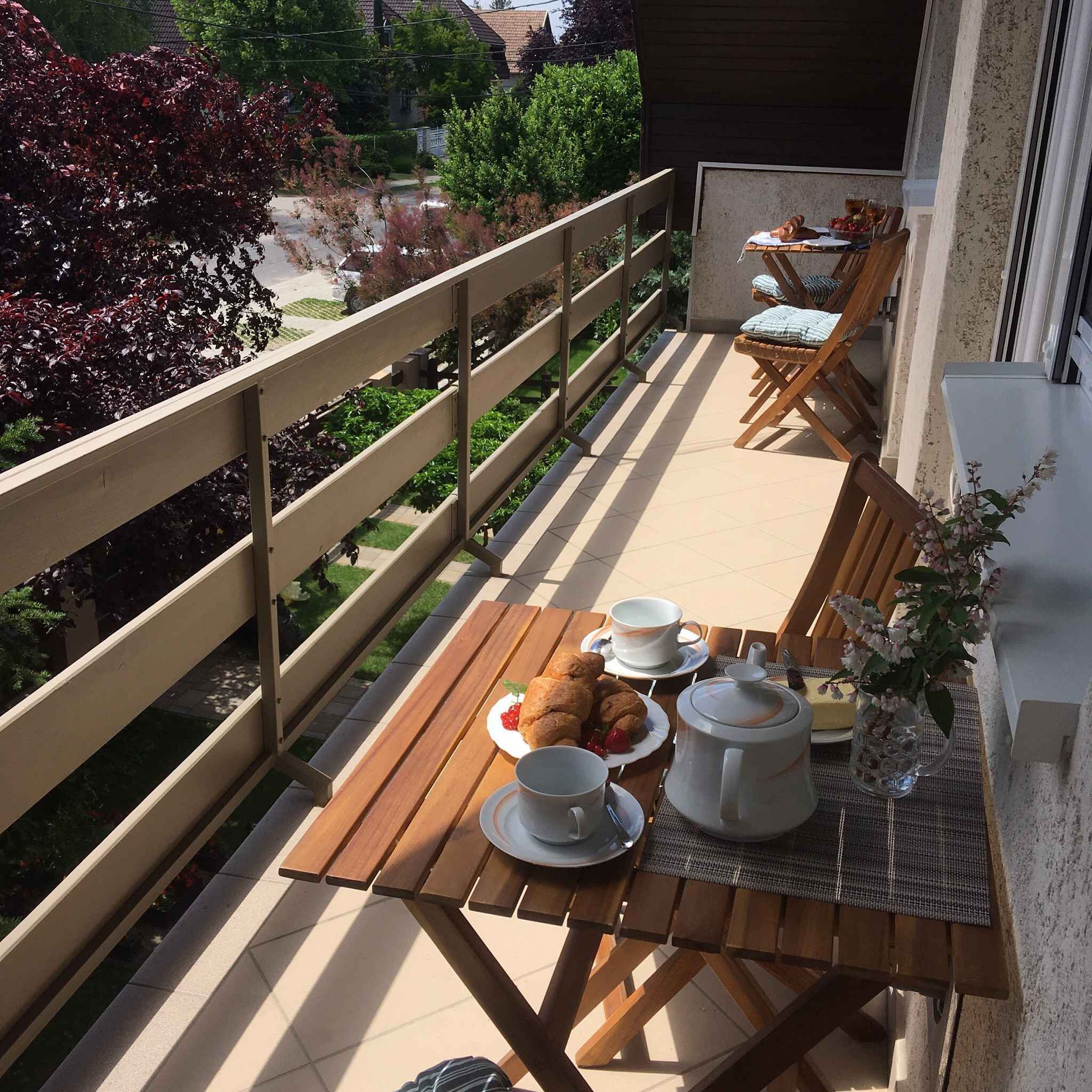 Ein Holztisch mit Frühstücksaufteilung auf einem Balkon, darunter Croissants, eine Kaffeekanne, Tassen und eine Vase mit Blumen.