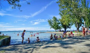 Eine Strandansicht mit badenden und spielenden Menschen, einem sandigen Ufer und Bäumen, die Schatten spenden.