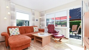 Ein modernes Wohnzimmer mit Sofa, Sessel und Hocker, einem Holztisch und Blick ins Freie durch große Fenster.
