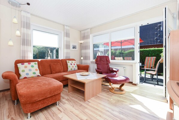 Un salon moderne comprenant un canapé, un fauteuil et un pouf, une table basse en bois, et une vue sur l’extérieur grâce à de grandes fenêtres.