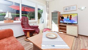 Ein modernes Wohnzimmer mit Fernseher, einem roten Sessel, einem Holztisch und Blick auf einen Garten durch große Fenster.