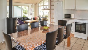 Uma área de refeições com uma mesa estampada, cadeiras pretas e uma tigela de frutas. Cozinha visível ao fundo, com micro-ondas e forno.