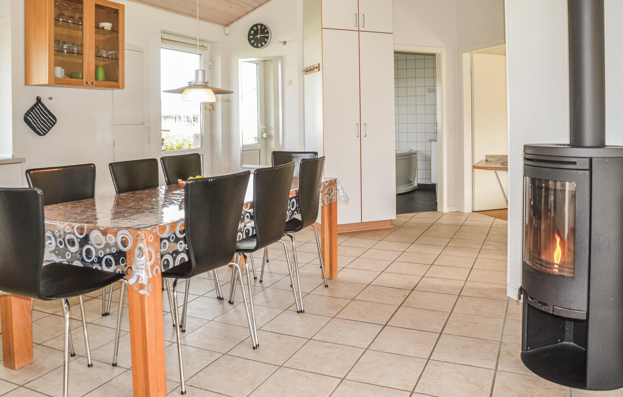 Een eetgedeelte met een houten tafel en stoelen, een fornuis, en een keuken met kasten en een klok.