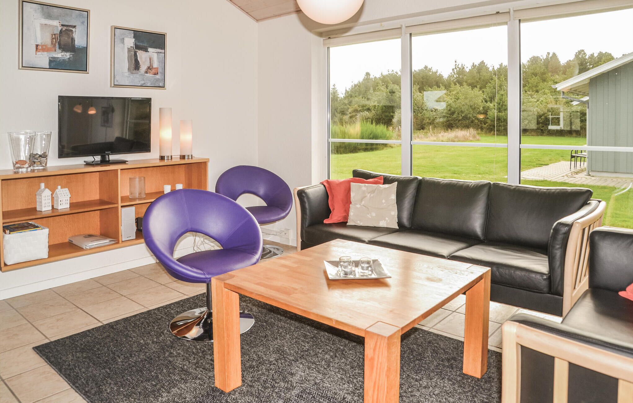 Un salon moderne avec une table basse en bois, deux fauteuils violets, un canapé en cuir noir et un téléviseur posé sur un meuble en bois.