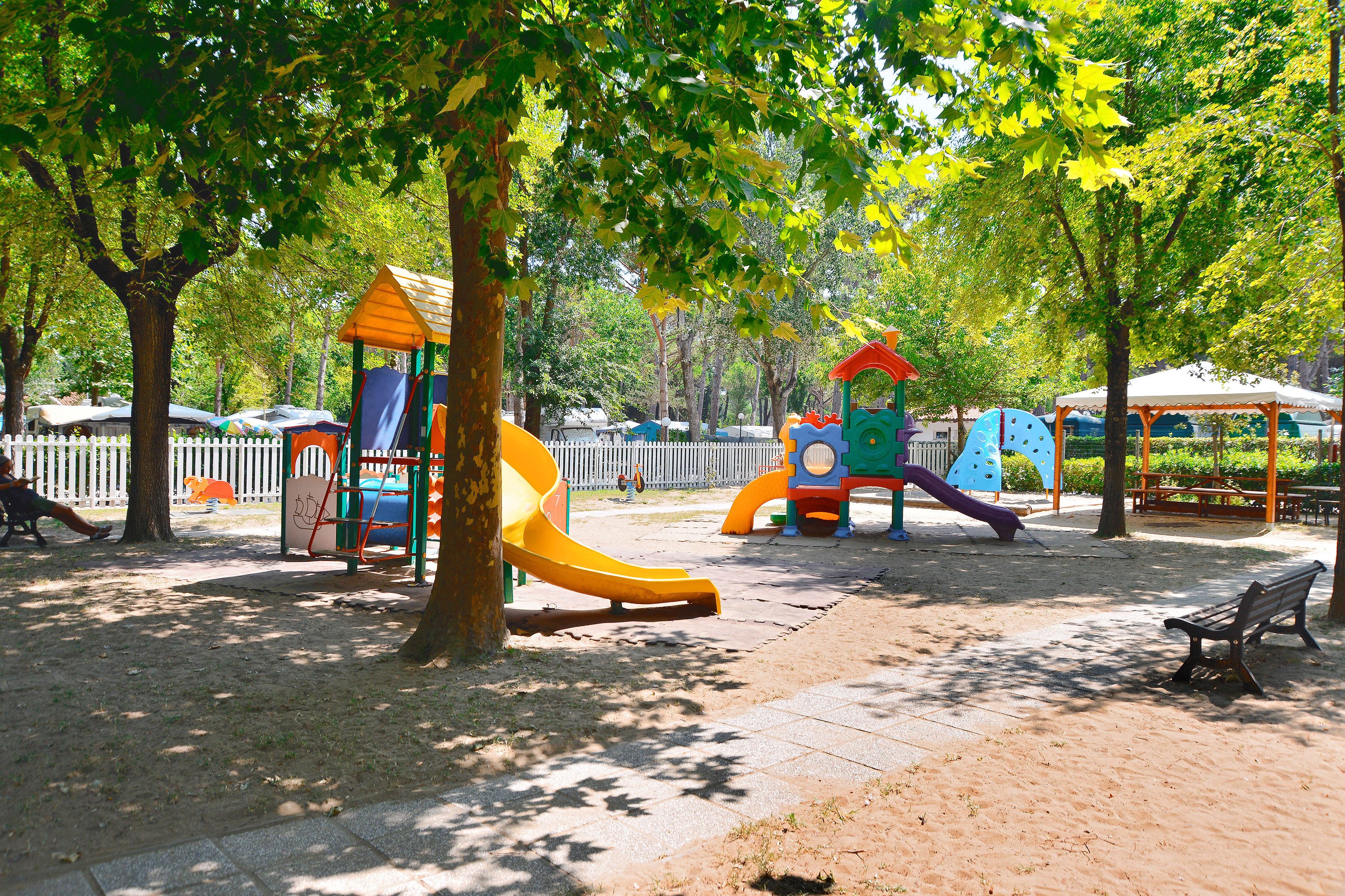 A playground with slides and climbing structures, surrounded by trees and a white fence.