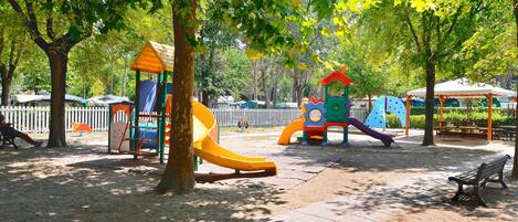A playground with slides and climbing structures, surrounded by trees and a white fence.