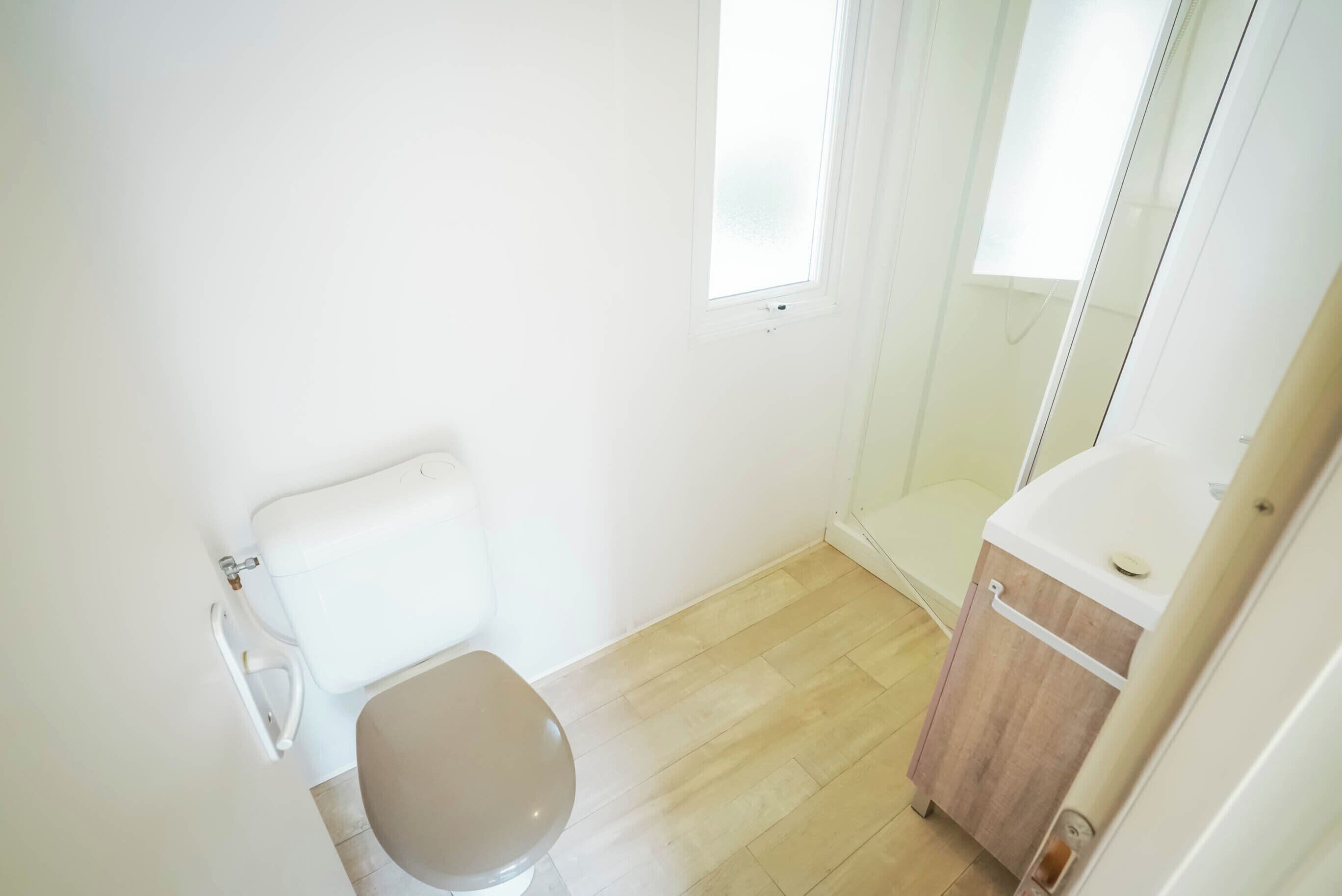 Ein kleines, helles Badezimmer mit Toilette, einem Holzschrank und einer Duschkabine aus Glas.