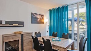 A dining area with a wooden table, black chairs, a small wooden cabinet, a painting, and a view through a glass door with blue curtains. - Villa Maria - Gabriele Whg. 13 - Maria 13 (Loddin)