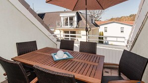 A terrace with a wooden table and wicker chairs, an umbrella, and a view of buildings in the background. - Villa Maria - Gabriele Whg. 13 - Maria 13 (Loddin)