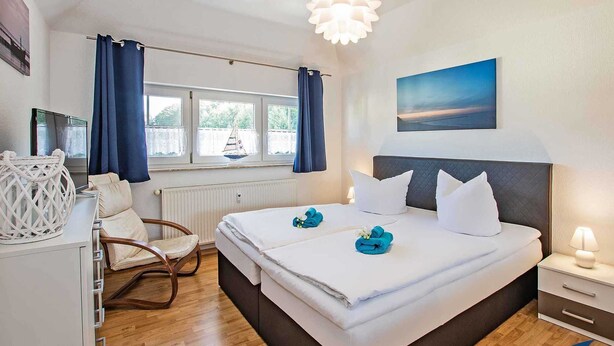 A hotel room with a bed, a chair, a desk, a window with curtains, a radiator, and a painting on the wall. - Villa Maria - Gabriele Whg. 13 - Maria 13 (Loddin)