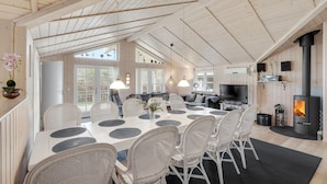 Ein geräumiges Zimmer mit hohen Decken, einem Essbereich mit weißen Korbstühlen und einem runden Tisch, einer Küche mit Herd und großen Fenstern, die viel Tageslicht hereinlassen.