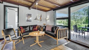 Ein modernes Wohnzimmer mit einer schwarzen Ledersofa, Holzstühlen und einem runden Couchtisch. An der Wand befinden sich abstrakte Gemälde und ein großes Fenster mit Blick auf Grünflächen.