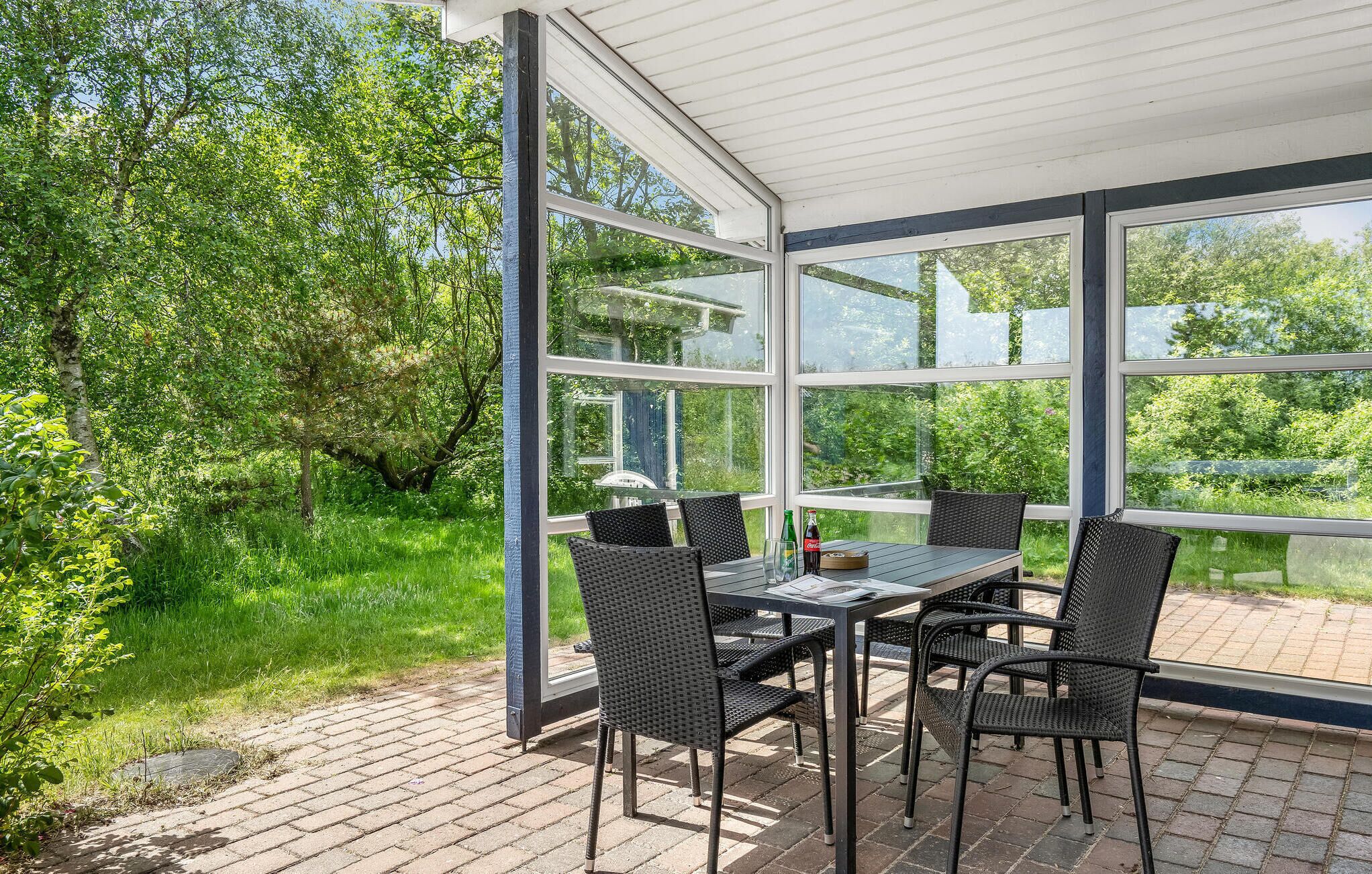 A covered patio with a table and chairs, surrounded by glass walls offering views of a lush garden.
