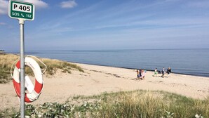 Una playa con un flotador sujetado a un poste, un cartel que indica 'P 405 SOS 112', y gente caminando por la orilla.