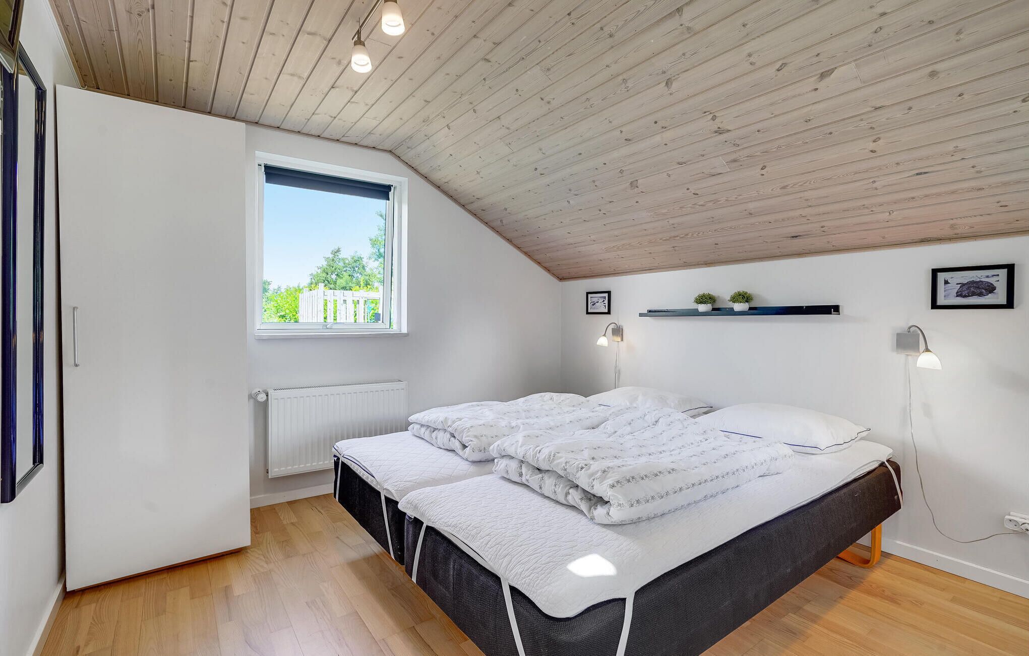 A bedroom with a sloped ceiling, a window with a view of greenery, a bed with white bedding, a radiator, and two wall-mounted lights.