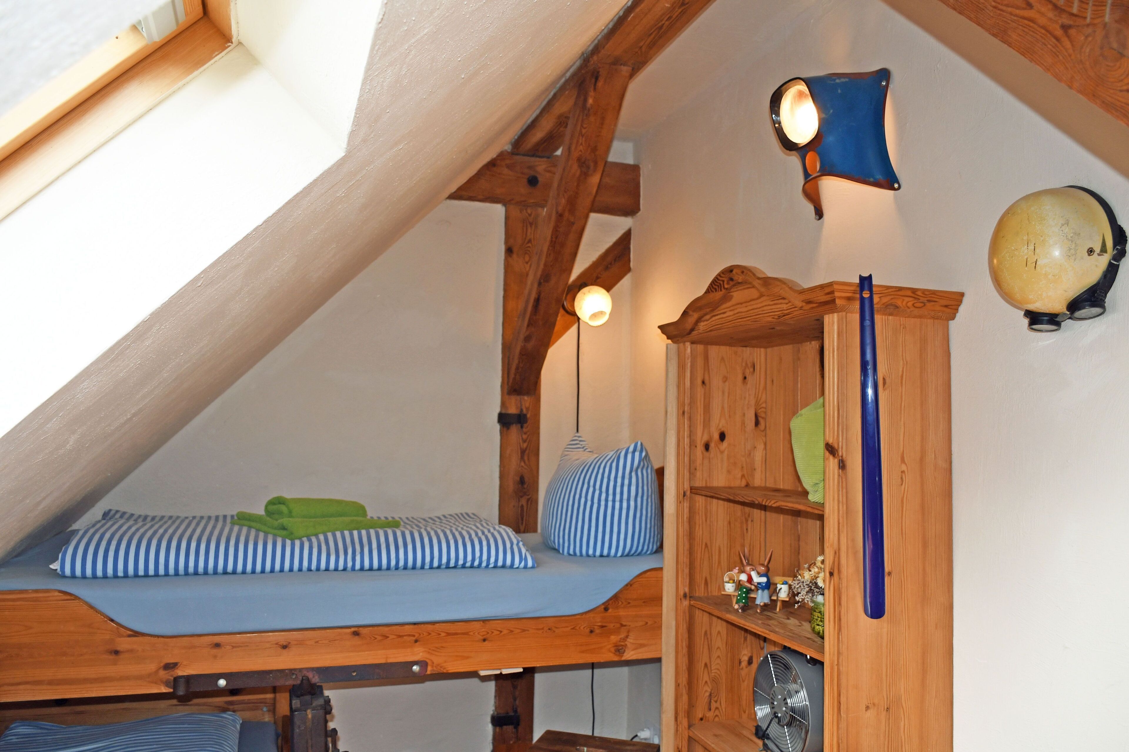 Une chambre à coucher aménagée dans un espace mansardé, avec un plafond en pente, des poutres en bois, un lit superposé, une petite étagère ornée d’objets décoratifs et un globe terrestre fixé au mur.