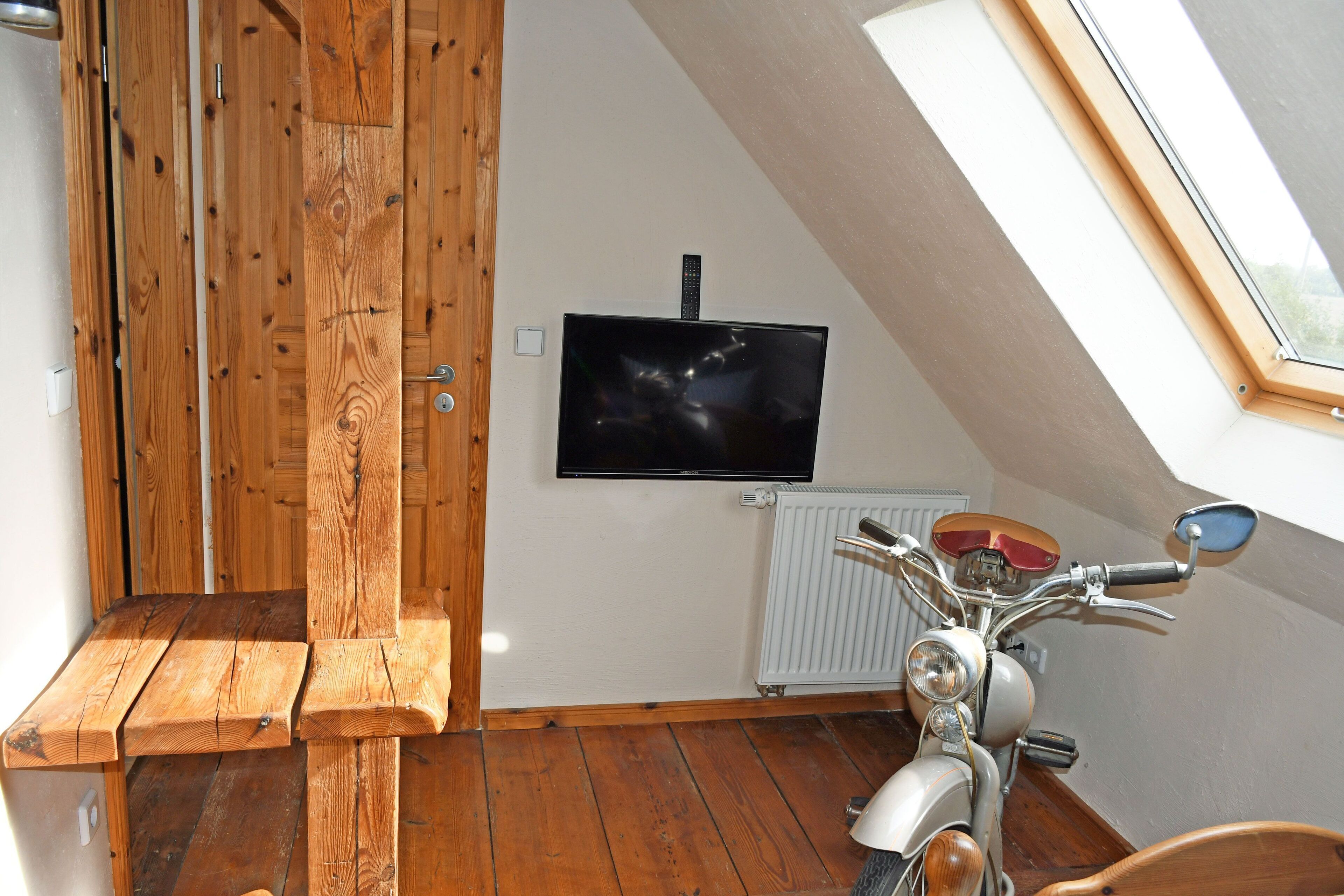 A room with a wooden bench, a motorcycle, a television mounted on the wall, and a skylight.