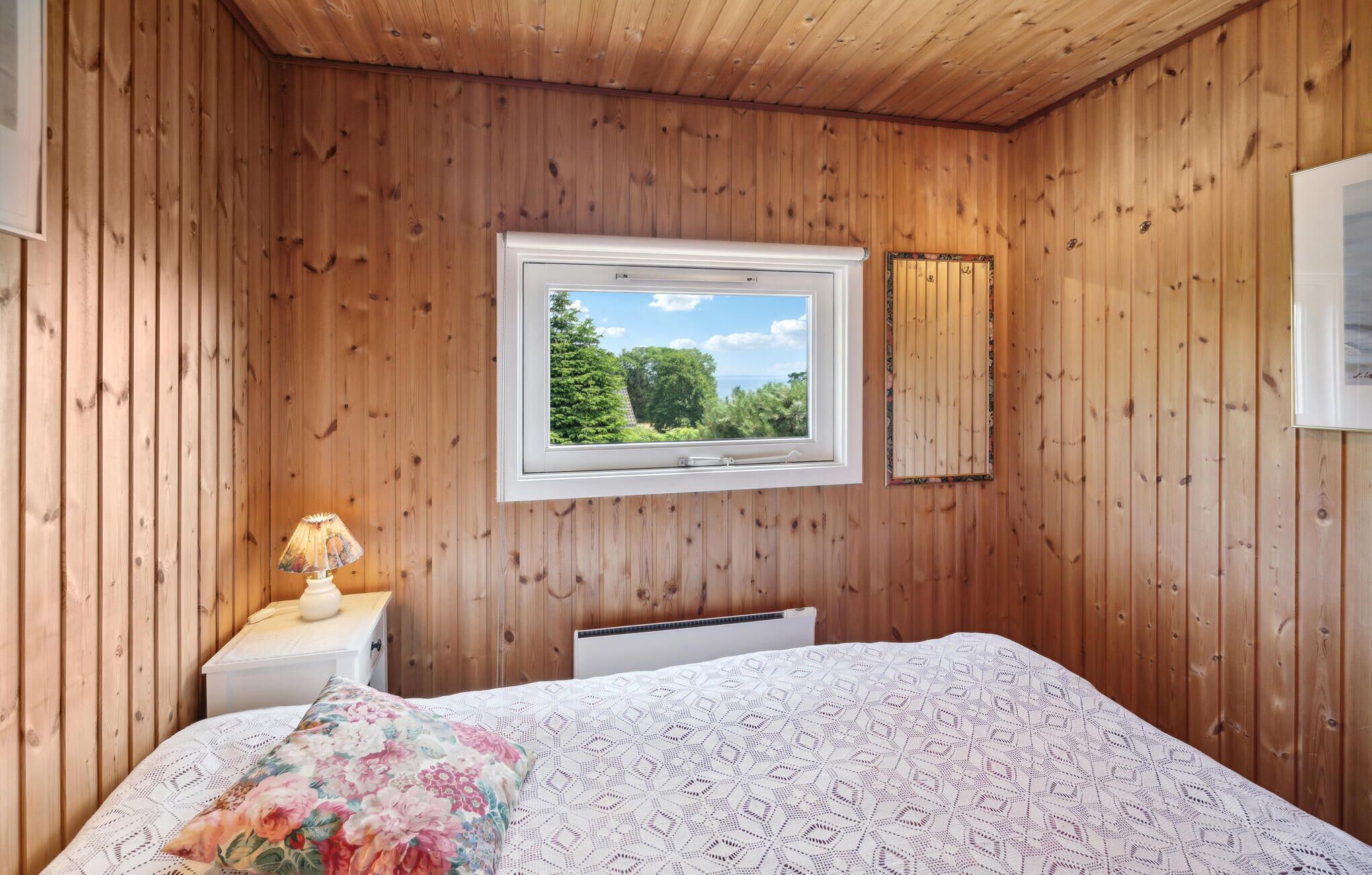 Une chambre avec des murs en bois, un lit orné d’un coussin à motifs floraux, une table de chevet avec une lampe, et une fenêtre donnant sur des arbres et le ciel.