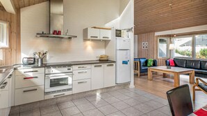 Moderni keittiö, jossa on valkoiset kaapit, sisäänrakennettu uuni ja jääkaappi. Keittiössä on myös ruokailutila, jossa on puinen pöytä ja tuolit, sekä sohva koristetyynyineen.
