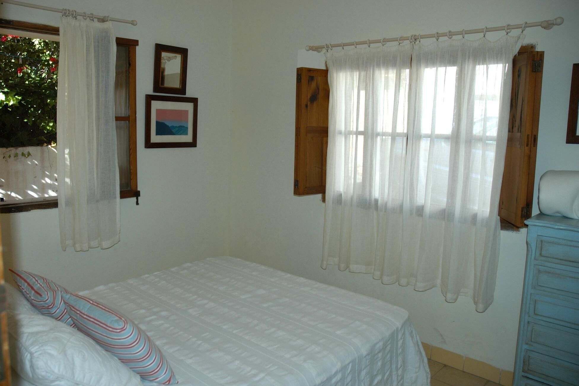 A bedroom with a bed, white curtains, a wooden wardrobe, and two framed pictures on the wall.