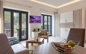 A modern living room with a sofa, a dining table with a bowl of fruit, a chair, and a view of the outdoors through large glass doors.