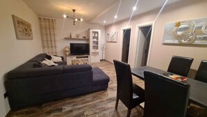 A modern living room with a dark sofa, a dining table with chairs, a flat-screen TV, and a ceiling light fixture. - Holiday apartment with garden terrace near the beach Špadići Poreč (Poreč)