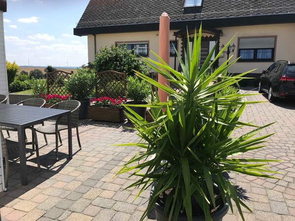 Een terras met een tafel en stoelen, plantenpotten en een gebouw met schuine dak in de achtergrond.