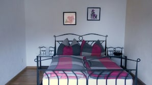 Een eenpersoonsbed met een metalen frame, een geruit sprei en meerdere kussens. Boven het bed hangen twee ingelijste foto’s.