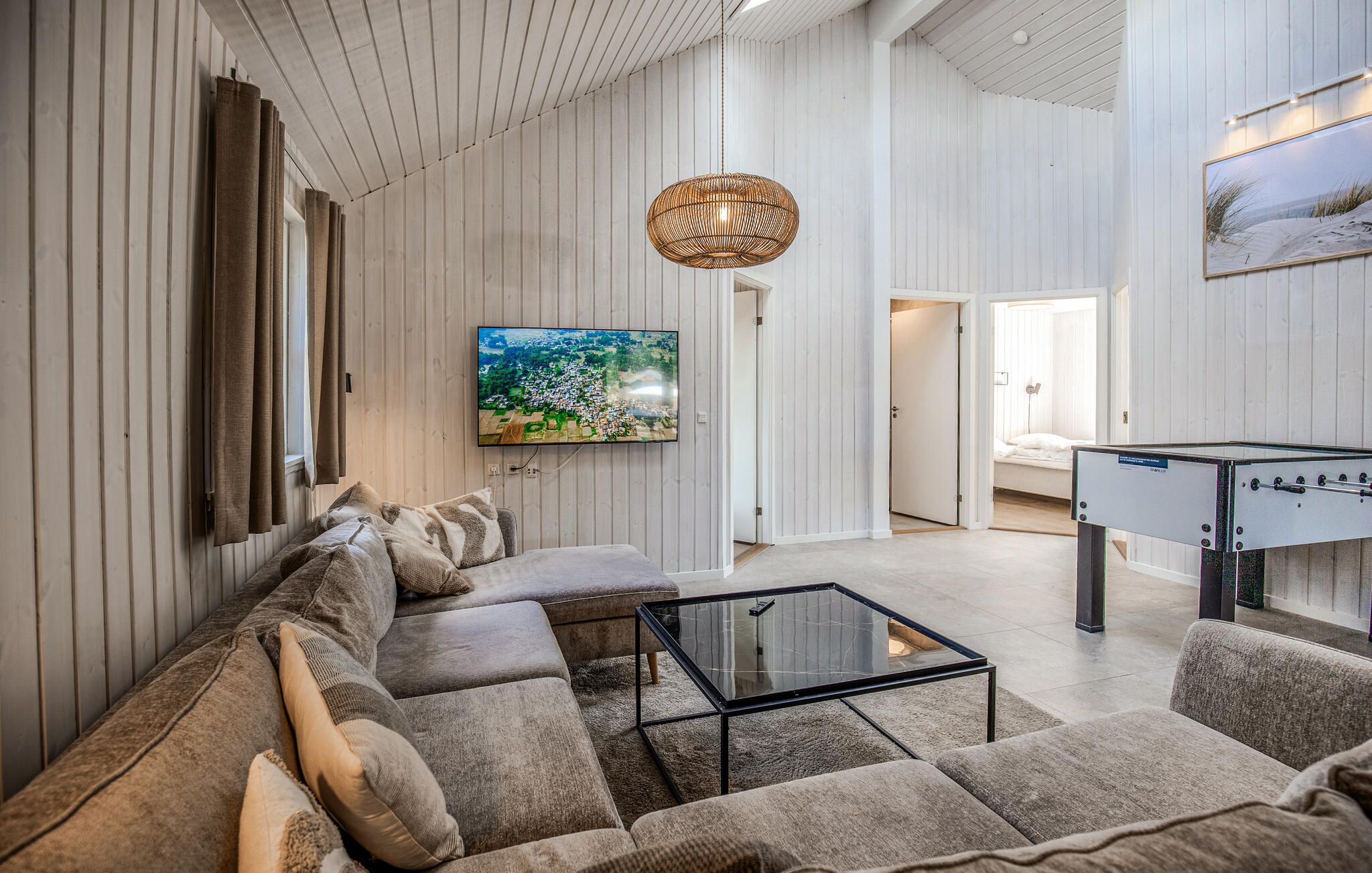 Een moderne woonkamer met een bank, een glazen salontafel, een aan de muur bevestigde televisie en een hanglamp.