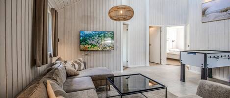 Een moderne woonkamer met een bank, een glazen salontafel, een aan de muur bevestigde televisie en een hanglamp.