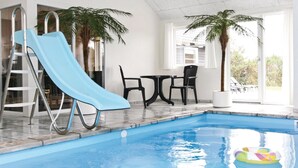 An indoor swimming pool with a blue slide, a small table, two chairs, and a potted palm tree. - Beautiful home in Vejers Strand with sauna (Vejers Strand)