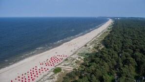 Eine Vogelperspektive zeigt einen Strand mit einer weiten Sandfläche, bewaldetem Gebiet und dem Meer.