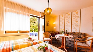 A room with two beds, a sofa, a table with fruits, and a view of greenery outside. - Vacation apartment 1 - Family-run guesthouse & vacation apartments Fischer-Haus (Trassenheide)