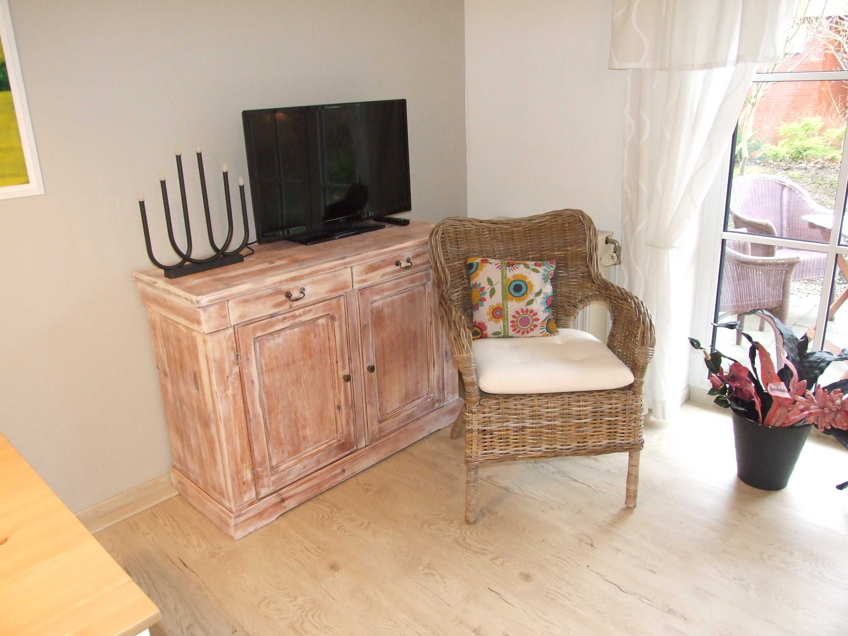 A room with a wooden cabinet, a wicker chair with a cushion, a television on the cabinet, and a window with curtains.