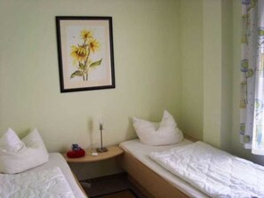 A room with two single beds, a bedside table with a lamp, a framed picture of sunflowers on the wall, and a window with a curtain. - Apartment Spilgies GM 69756 - Apartment Spilgies SNR: 83 (Graal-Müritz)