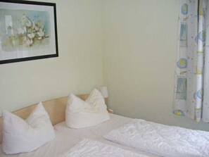 A single bed with white bedding and pillows, a framed picture on the wall, and a curtain with a pattern.